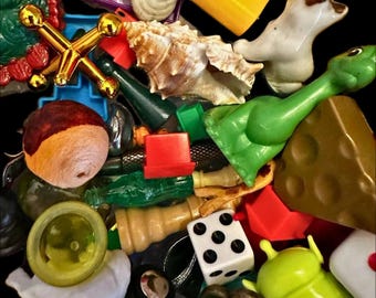 May include: A collection of assorted small objects including a gold-colored metal jack, a white seashell, a green plastic dinosaur, a white die with black dots, a brown wedge of cheese, a wooden sphere, and various other colorful plastic trinkets.