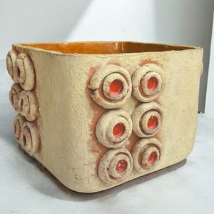 May include: A square ceramic planter with a textured surface and a brown rim. The planter has a pattern of raised circles with red centres.
