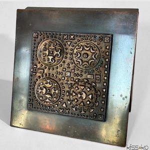 May include: A square, metal decorative piece with a central embossed design. The design features four circular patterns with intricate details, surrounded by a textured border. The metal has a tarnished, aged appearance.