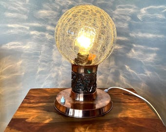 May include: A copper table lamp with a textured glass globe shade. The shade has a crackled finish and a light bulb is visible inside. The lamp base has a decorative pattern.