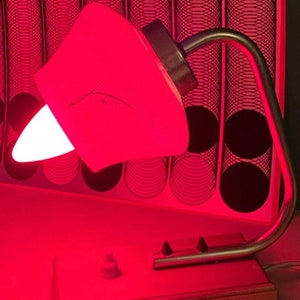 May include: A vintage desk lamp with a white shade and a silver metal base. The lamp is turned on and casting a red glow.