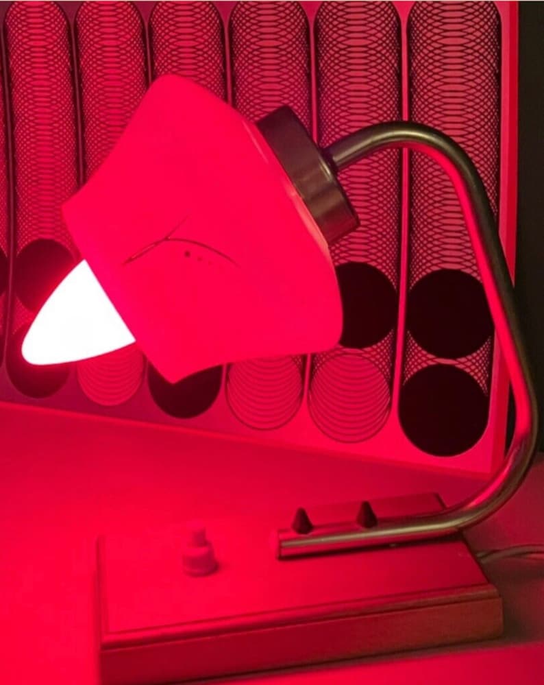 May include: A vintage desk lamp with a white shade and a silver metal base. The lamp is turned on and casting a red glow.