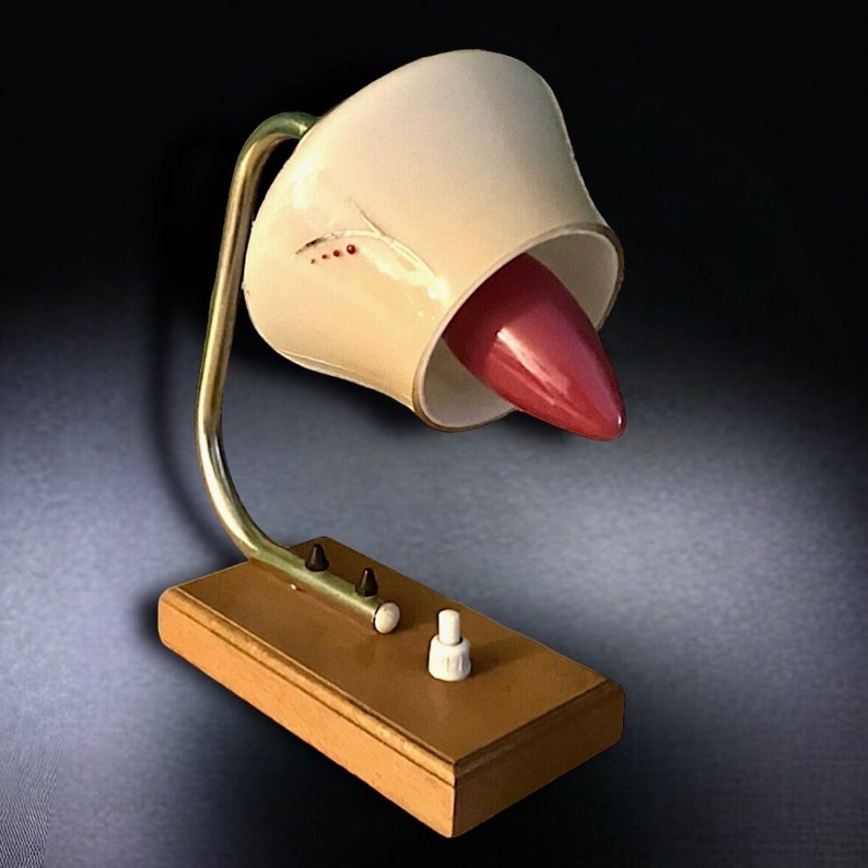 May include: A vintage table lamp with a cream-colored shade and a pink accent. The lamp has a gold-colored metal base and a wooden base.