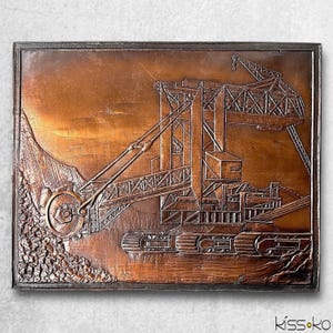 May include: A copper-toned relief sculpture of a large industrial excavator, set within a dark brown frame. The artwork showcases detailed carvings of the machine's components, including a large wheel, tracks, and a boom. The background suggests a landscape.