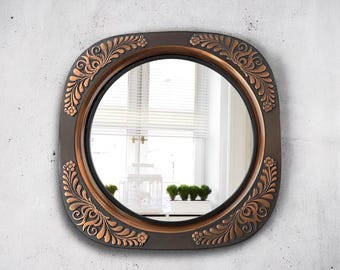 May include: A round mirror with a square frame. The frame is dark brown with copper-colored floral and leaf designs. The mirror reflects a window with blinds and a white birdcage.
