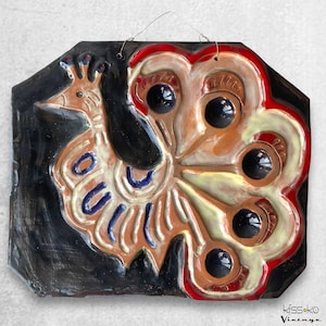 May include: A ceramic wall hanging featuring a stylized peacock design. The artwork showcases a black background with a colorful peacock in shades of brown, cream, red, and blue. The piece has a wire for hanging and the text "kissko Vintage".