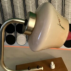 May include: A vintage table lamp with a white glass shade featuring a floral design. The lamp has a silver metal arm and base with a wooden base. The lamp has a white switch.