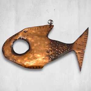 May include: A copper fish-shaped wall hanging with a large circular opening and a pointed tail. The fish has a textured surface with small, evenly spaced dots and a loop for hanging. The metal has a warm, reddish-brown hue.