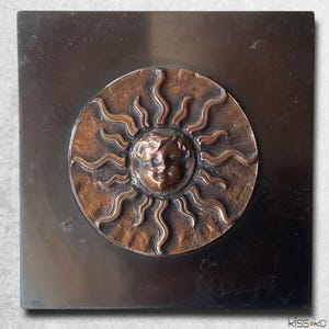 May include: A square, dark metal plaque with a circular, copper-toned sunburst design. The sun has a smiling face at the center, with wavy rays extending outward. The plaque has a brushed metal finish.