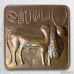 May include: A square, bronze-coloured relief sculpture featuring a stylised bull with large horns and a decorative collar. The relief includes ancient symbols and text above the bull, with a wheel-like symbol on the left. The artwork has a vintage aesthetic.