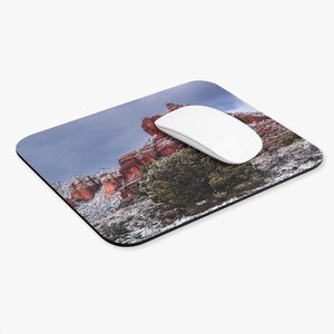 Pode incluir: Um mousepad retangular com uma paisagem de formações rochosas vermelhas cobertas de neve sob um céu nublado. Um mouse de computador branco repousa sobre o mousepad. O mousepad tem uma borda preta.