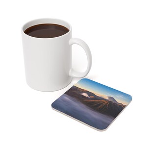 May include: A white ceramic mug filled with dark liquid, next to a square coaster. The coaster features a landscape image of a mountain range with a blue sky and clouds. The mug has a curved handle.