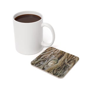 May include: A white ceramic mug filled with dark liquid, next to a square coaster. The coaster features a detailed image of a Buddha head nestled within tree roots, in shades of brown and tan.