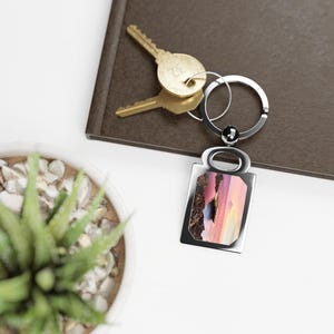 May include: A rectangular silver keychain with a colorful landscape image. The keychain is attached to a silver ring with two gold keys. A small succulent plant in a white pot is in the foreground.
