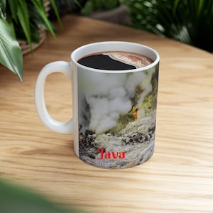 Peut inclure: Mug en céramique blanche remplie de café noir. La tasse présente une image panoramique d'un paysage volcanique avec de la vapeur et le mot "Java" en rouge. La tasse est posée sur une surface en bois.