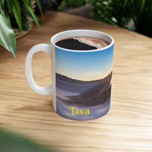 May include: White ceramic coffee mug filled with dark coffee. The mug features a scenic landscape design with mountains and a blue sky, and the word "Java" in yellow.
