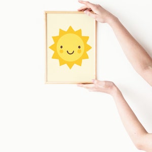 Puede incluir: Una impresi&oacute;n enmarcada de un sol amarillo de dibujos animados con una cara sonriente. El sol tiene un fondo amarillo claro y est&aacute; enmarcado en un marco de madera claro.