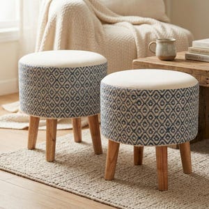 Può includere: Due sgabelli rotondi con sedute imbottite color crema e lati decorati con un motivo geometrico a rombi blu. Ogni sgabello è sostenuto da quattro gambe in legno di tonalità chiara, che offrono una base stabile per questi mobili funzionali.