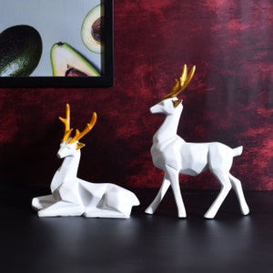 May include: Two white ceramic deer sculptures with gold antlers. One deer is standing and the other is lying down.