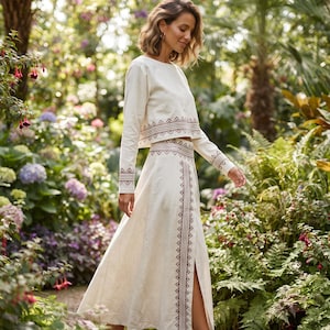 Op de afbeelding: Een crèmekleurige tweedelige set bestaande uit een cropped top met lange mouwen en een maxi-rok met hoge taille. Beide kledingstukken zijn voorzien van bruin geometrisch borduurwerk langs de zomen en een verticale decoratieve streep.