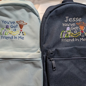 Op de afbeelding: Twee blauwe en zwarte rugzakken met geborduurde afbeeldingen van Woody en Buzz Lightyear uit Toy Story. De tekst op de rugzakken luidt "You've Got a Friend in Me" en "Jesse You've Got a Friend in Me".