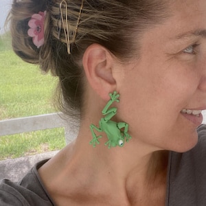 May include: A pair of light green frog earrings. The earrings are a whimsical design, with the frog's legs and arms extended. Each frog has a single, clear gem for an eye. The earrings are worn on a person.