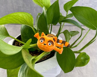 May include: A small orange and black cat figurine with a surprised expression, sitting on a green potted plant.