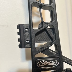 May include: Black compound bow with a black and silver "Mathews" logo on the riser. The bow has a black accessory mount on the side.