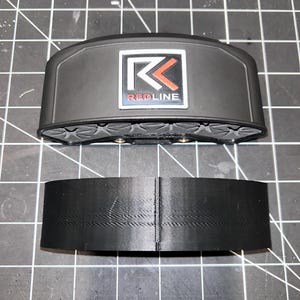 May include: A black plastic 3D printed part with a red and white logo that says "REDLINE" and a white letter "K" inside a square. The part is designed to be attached to a larger object.