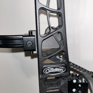 May include: Close-up of a black compound bow with the brand name "Mathews" visible. The bow features a cut-out design on the riser and a black stabilizer bar. The bow is set against a plain white background.