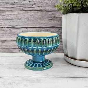 May include: A teal-colored, footed bowl with a ribbed design. The bowl has a wide, open top and a pedestal base. The interior is a light cream color. The bowl is likely ceramic or glass and is displayed on a white wooden surface.