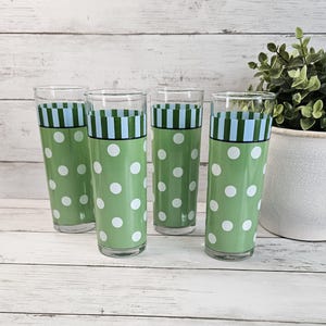 May include: A set of four tall, cylindrical clear glass tumblers featuring a vibrant green base decorated with white polka dots. Each glass is accented with a horizontal band of light blue and dark green vertical stripes near the top rim.