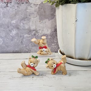 May include: Three ceramic teddy bear figurines with red bows and holly accents. One bear is upside down, another is lying down, and the third is standing. The bears are tan with black eyes and are on a white wooden surface.