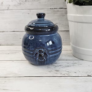 May include: A round, dark blue ceramic jar with a matching lid. The jar features a glossy, speckled glaze and a raised, molded bee emblem on the front. The lid has a small, rounded knob handle at the top, and the body has horizontal ridges.