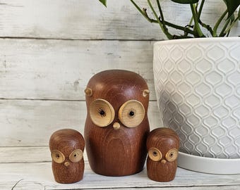 以下が含まれることがあります： 木製のフクロウの置物3個セット。一番大きなフクロウは茶色で、大きく丸い目があります。2つの小さなフクロウも茶色で、目は小さくなっています。フクロウは白い木製の表面に一列に並んでいます。