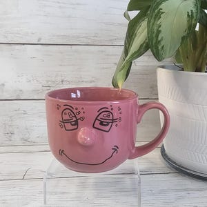May include: A dusty rose ceramic mug featuring a raised, rounded nose in the center and a black line-art graphic of two sleepy, half-closed eyes with eyelashes and a wide, curved smile. The mug has a single handle on the right side.
