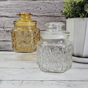 May include: Two glass jars with lids. One jar is clear, and the other is amber-colored. Both jars have a square shape with embossed floral designs. The jars are on a white wooden surface, with a plant in a white pot in the background.