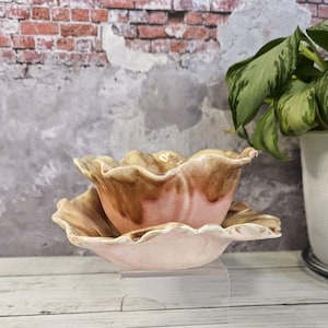 May include: A set of two ceramic bowls with a ruffled edge. The bowls are a light pink color with brown and beige glaze. The bowls are stacked on top of each other.