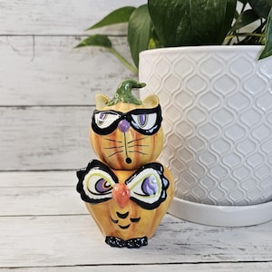 May include: A stacked ceramic Halloween decoration featuring a cat-like pumpkin design. The orange pumpkins have black-rimmed glasses and a green hat. The bottom pumpkin has owl-like features. The decoration is on a white wooden surface.