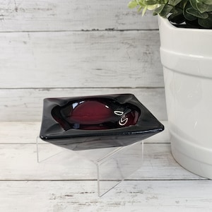 May include: A square, deep purple glass ashtray with a smooth, polished finish and four rounded indentations along the edges. The ashtray features a thick, translucent glass construction that catches the light, showcasing a rich, dark plum color throughout the bowl.