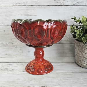 May include: A red and green ceramic pedestal bowl with a textured surface. The bowl is on a white wooden background.
