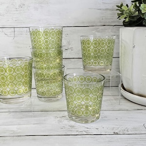 May include: A collection of six clear drinking glasses with a green floral pattern. The glasses are arranged on a clear acrylic surface, with a white ceramic pot and greenery in the background. The glasses vary in height.