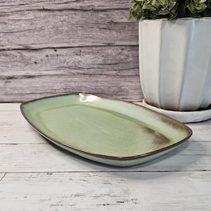May include: A rectangular, light green ceramic serving platter with a brown rim. The platter has a slightly textured surface and is displayed on a white wooden surface. A white, geometric planter with greenery is in the background.