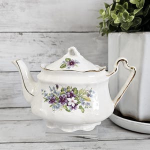 May include: A white porcelain teapot with a floral design featuring purple and white flowers and green leaves. The teapot has a gold trim and a matching lid. The handle is heart-shaped. The teapot is displayed on a clear stand.
