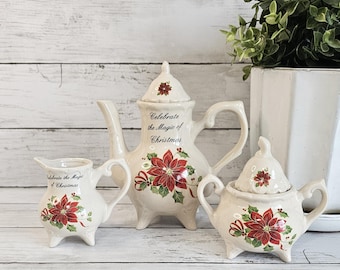 Peut inclure: Un service à thé de Noël crème, comprenant une théière, un pot à crème et un sucrier. Chaque pièce est décorée de poinsettias rouges, de feuilles vertes et du texte "Celebrate the Magic of Christmas."