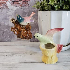May include: Two ceramic bird figurines. One bird is perched on a brown tree stump, with a blue, pink, and purple color scheme. The other bird is yellow, green, and red, with an open top, resembling a small vase.