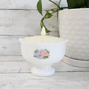 May include: A white, footed candy dish with a lid and a floral design. The dish features a pedestal base and a textured lid with a small, pointed finial. A cluster of pink and purple flowers decorates the front of the dish.
