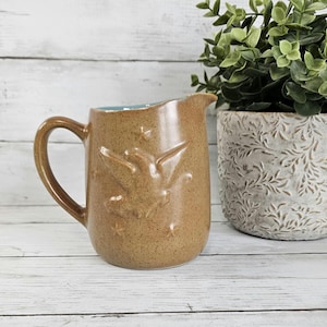 May include: A tan ceramic pitcher with a handle and a spout. The pitcher has a textured surface with embossed bird designs. The interior rim is light blue. A decorative plant in a white pot is in the background.