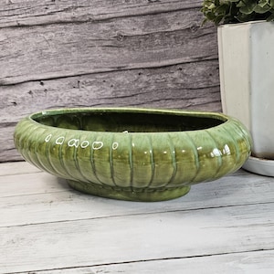 May include: An oval-shaped ceramic planter with a glossy, moss-green glaze and a ribbed, fluted exterior texture. The bowl features a wide, open top and a raised base, showcasing a deep green finish with subtle variations in color and tone.