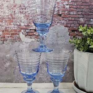 May include: Three blue-tinted glass goblets with a ribbed design. The goblets are stacked, with one on top and two below. The stems and bases are a lighter shade of blue. The background features a brick wall and a potted plant.